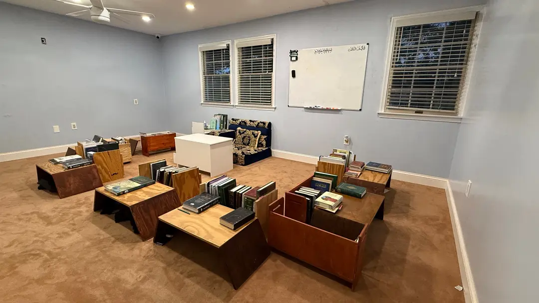 A classroom with pale blue walls: low wooden desks stacked with kitabs, a cushioned seat for the teacher, and a whiteboard headed in Arabic. The room is empty.