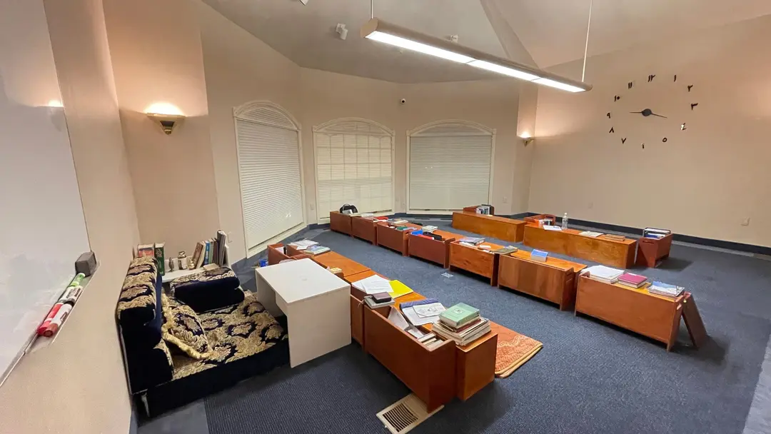 A high-ceilinged classroom with arched windows and a blue carpet: rows of long wooden desks with books and papers, and a wall clock with Arabic numerals. The room is empty.