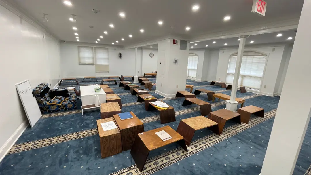 A large classroom with a blue patterned carpet and white columns: many low wooden desks set out across the floor, and a whiteboard leaning against the wall. The room is empty.