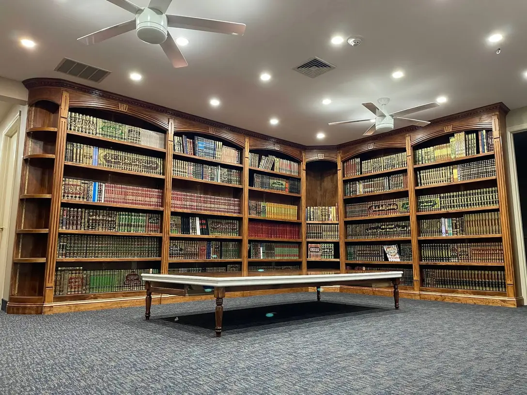 The Foundation’s library: arched wooden bookcases filling two walls, shelved floor to ceiling with bound kitabs, and a long low reading table on the carpet. The room is empty.