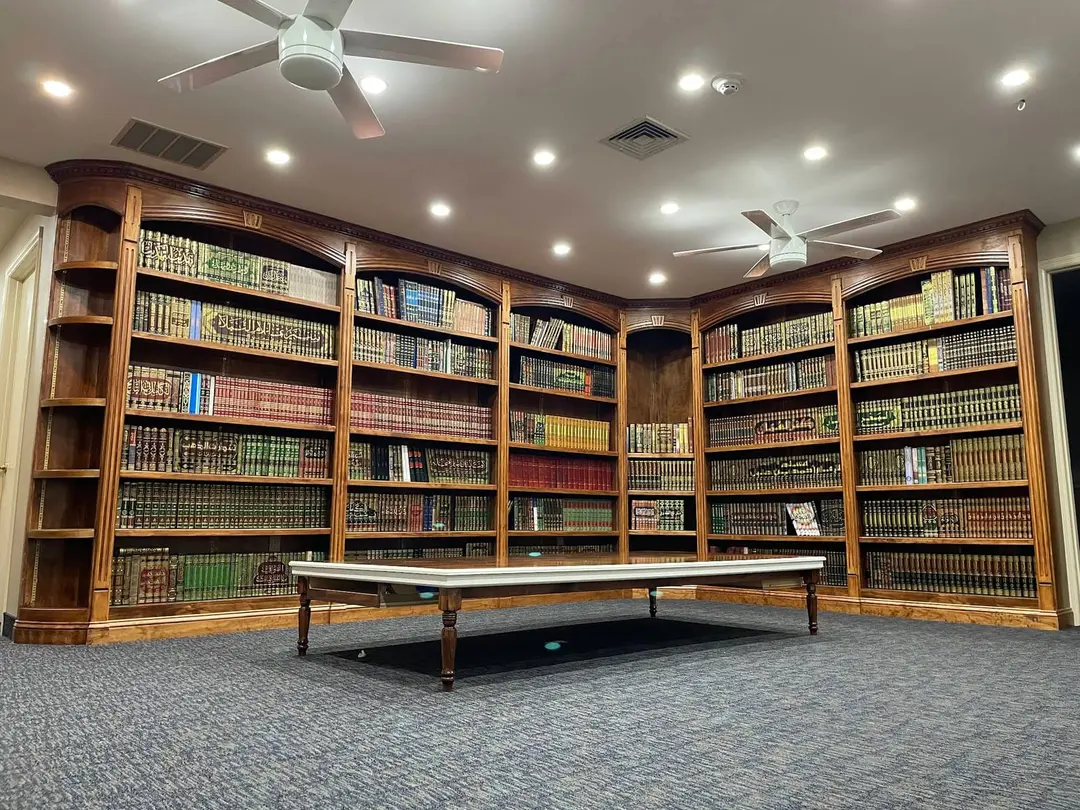The library: arched wooden bookcases filling two walls, shelved floor to ceiling with bound kitabs, and a long low reading table on the carpet. The room is empty.