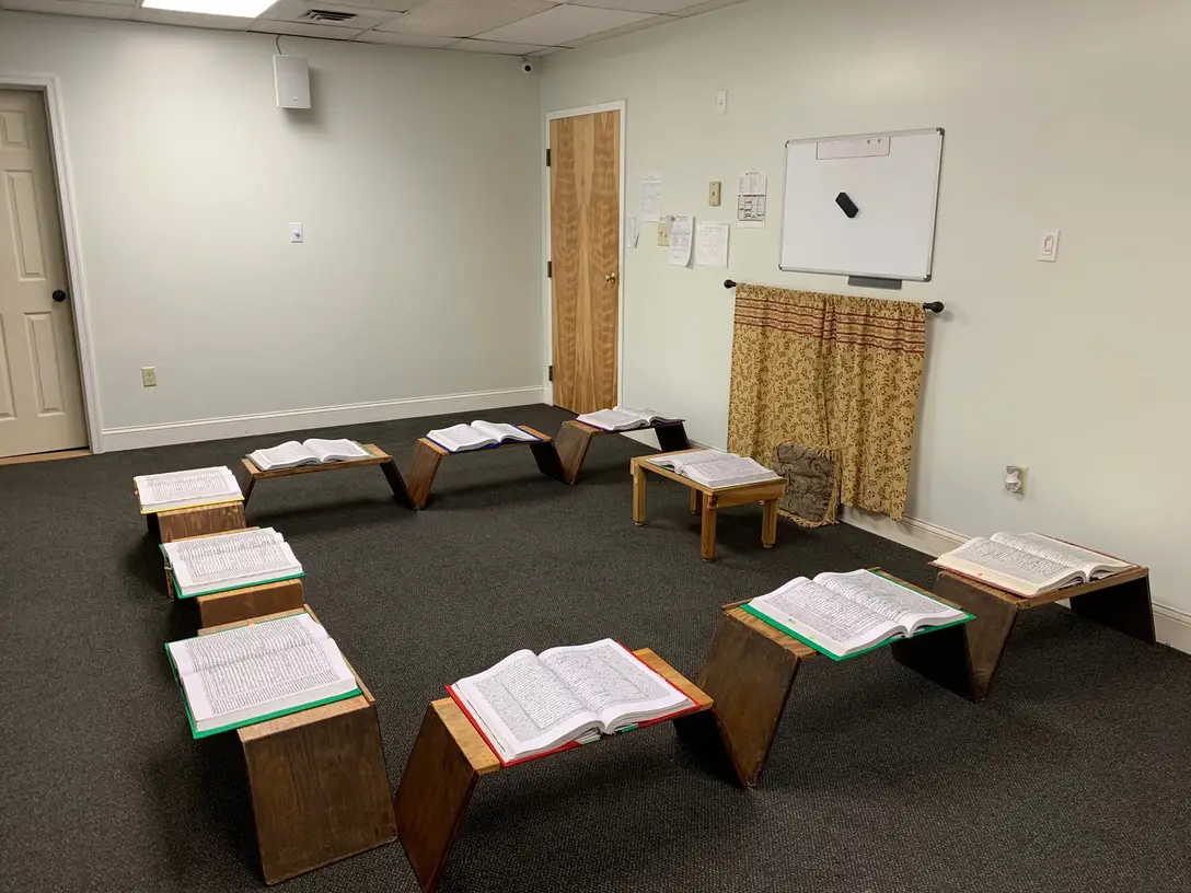 A classroom at the Foundation: low wooden rehals arranged in a horseshoe on the carpet, each holding an open kitab. The room is empty.