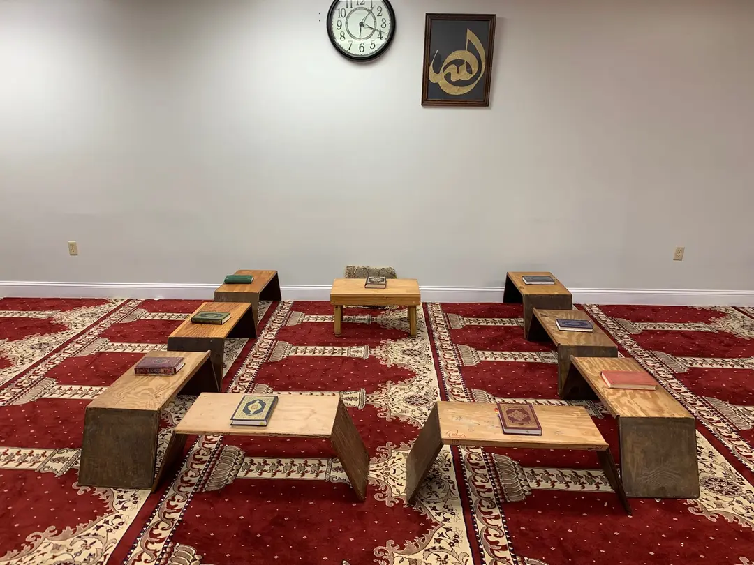 A Hifz classroom: wooden rehals set out on a red prayer carpet, each holding a closed Qur’an, with framed Arabic calligraphy on the wall. The room is empty.