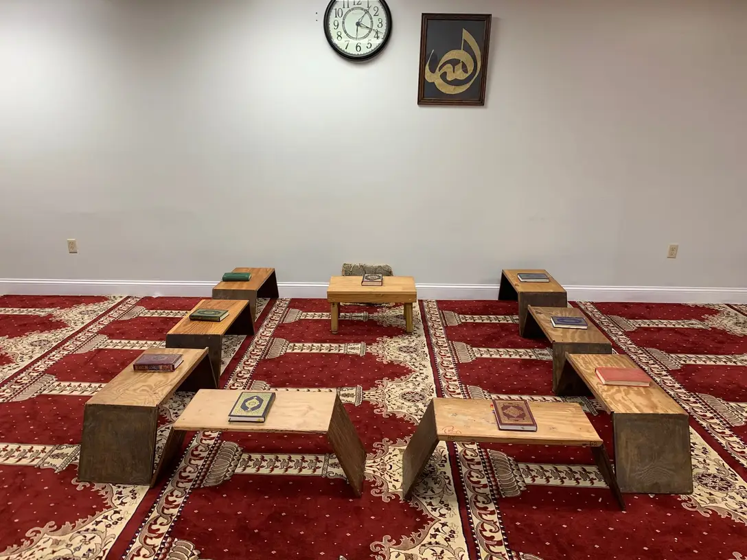 A Hifz classroom at the Foundation: wooden rehals set out on a red prayer carpet, each holding a closed Qur’an, with framed Arabic calligraphy on the wall. The room is empty.