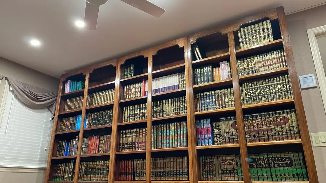 A second wall of the library: a tall wooden bookcase with scalloped tops, every shelf filled with bound sets of kitabs.