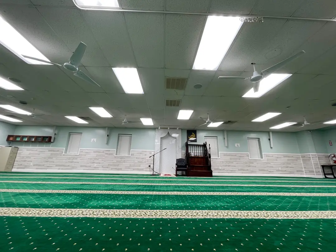The qibla wall of the Main Masjid seen low from the carpet: a white marble mihrab with a pointed arch, and a carved wooden minbar beside it.