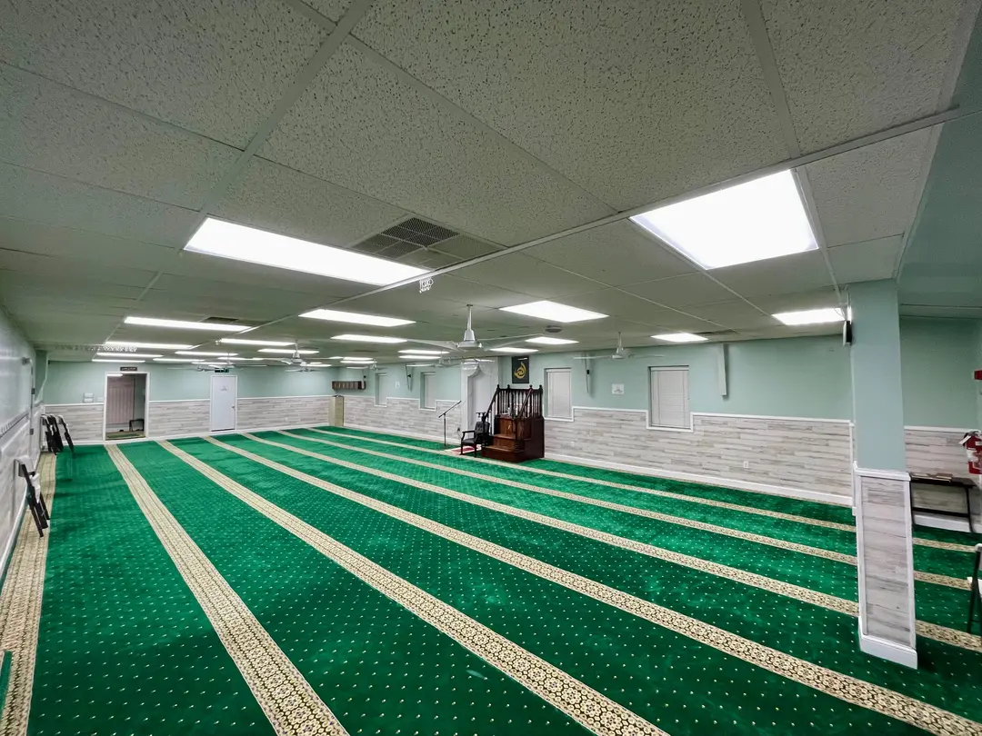 The prayer hall of the Main Masjid: green carpet marked in prayer rows running toward the far wall, where the minbar stands beside the mihrab. The hall is empty.