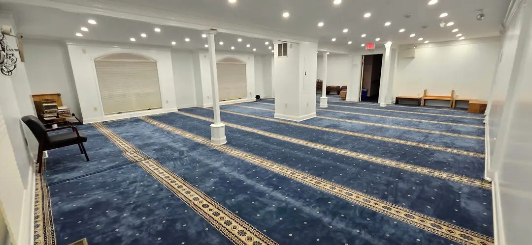 A wide musalla at the seminary: blue carpet marked in prayer rows, white columns, and arched windows along one wall. The room is empty.